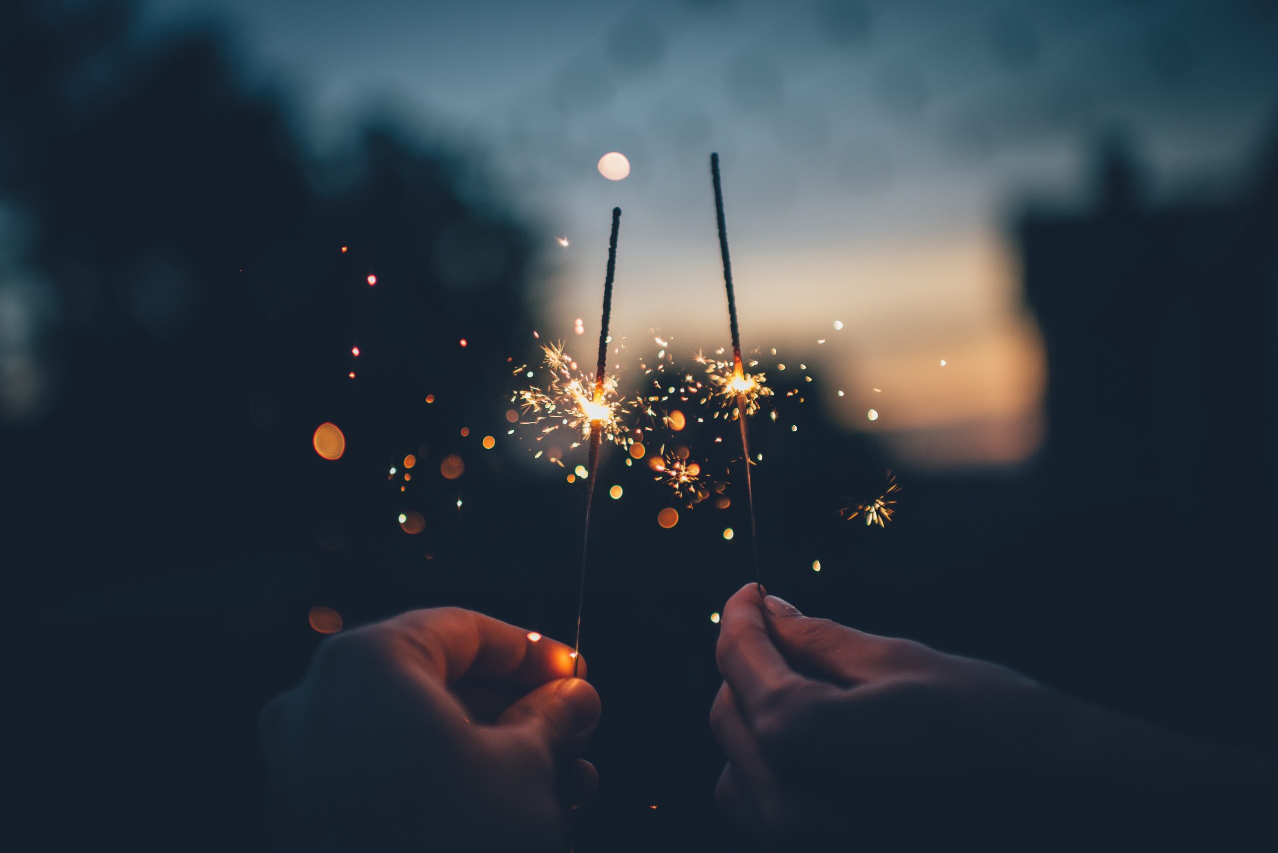 Depicts two hands holding sparklers. Used as a cover photo for a news article about year-end review emails from various alternative investing platforms.