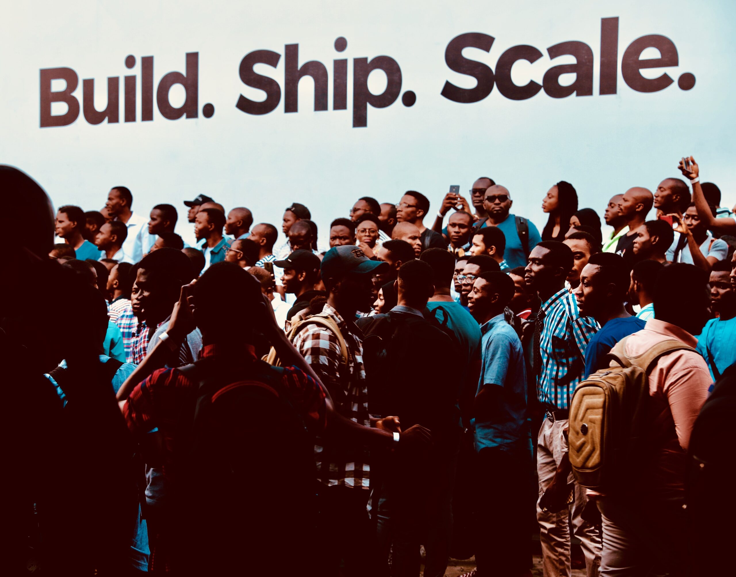 Depicts many people standing in front of a wall that says "Build. Ship. Scale." Used to represent startups / equity crowdfunding as an asset class.
