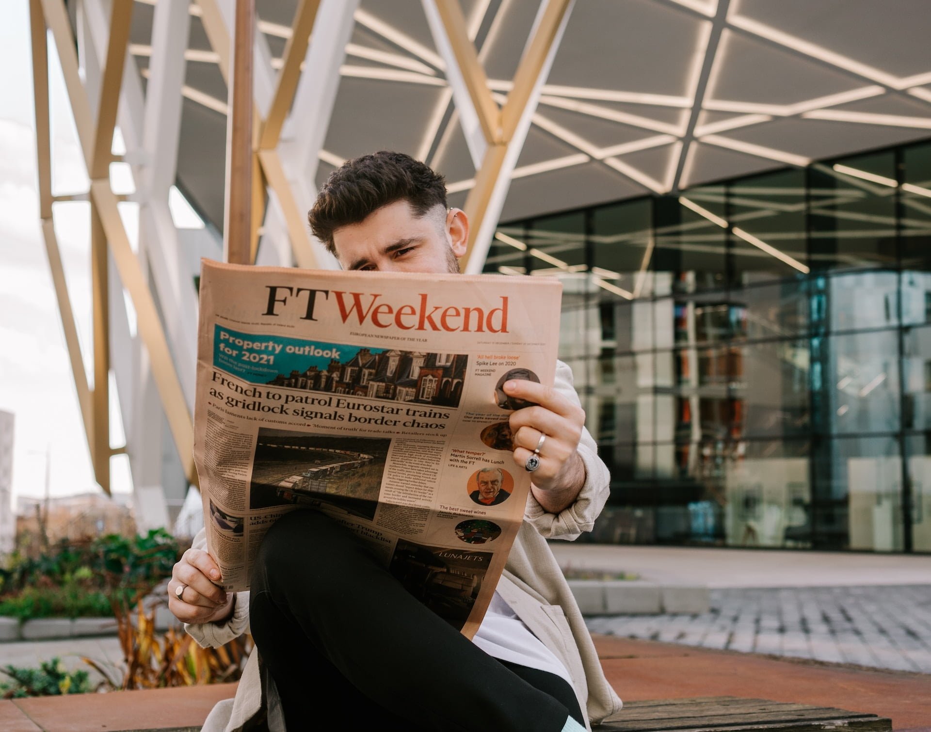 Depicts a man sitting outside and reading a financial newspaper. Used as a cover image for an article discussing alternative investment news in July.